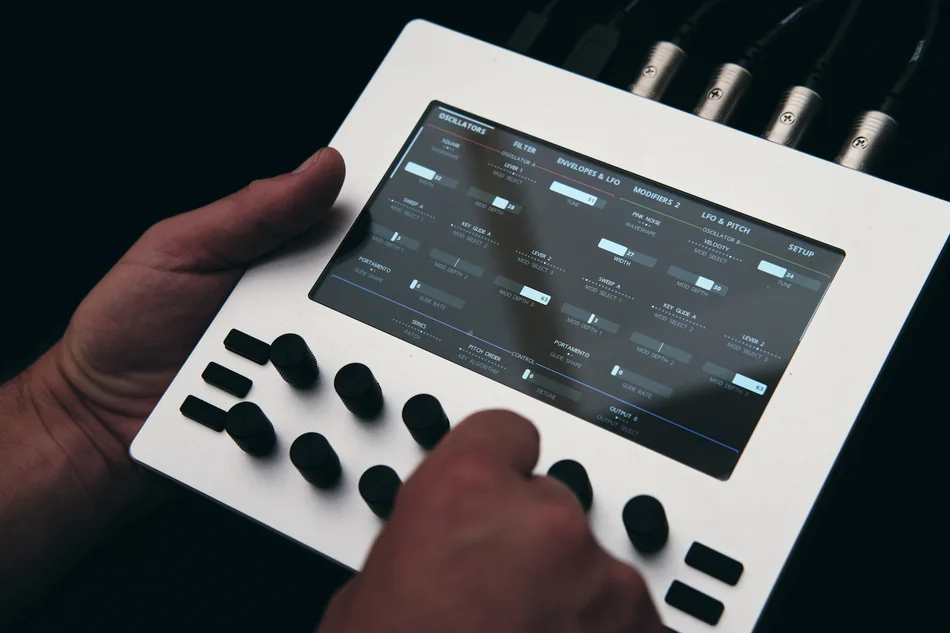 A white Electra One mk1 held in one hand, a thumb on its knobs and a page of synthesizer parameters lit on the screen