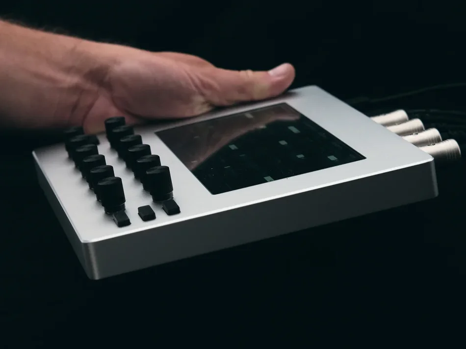 A hand resting on an Electra One mk1 against a black background, its MIDI cables running off to the right