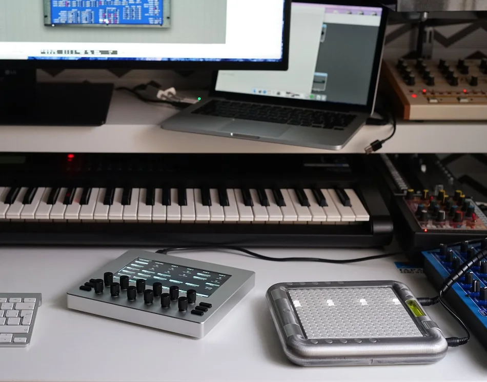 Electra One mk1 on a desk in front of a keyboard, a laptop and a rack of synthesizers