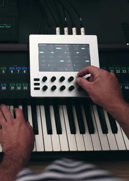 Hands playing a synthesizer keyboard with a white Electra One mk1 resting on it, showing a page of parameters