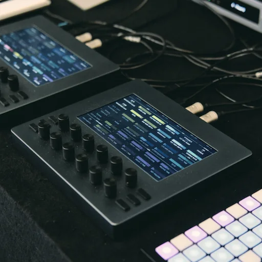 Two Electra One mk2 controllers on a table beside a grid pad controller, each showing a page of instrument parameters