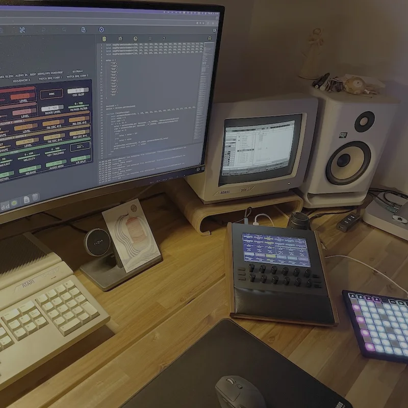 A studio desk: an Electra One mk2 on a wooden stand beside a grid pad
          controller, with a preset editor open on the monitor and an Atari ST
          and its monitor behind