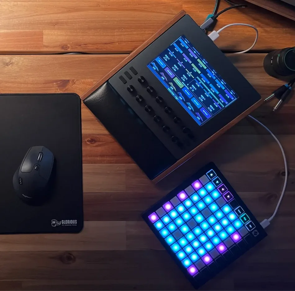 Electra One mk2 in a wooden stand on a desk, a grid pad controller plugged into it over USB with its pads lit blue and purple