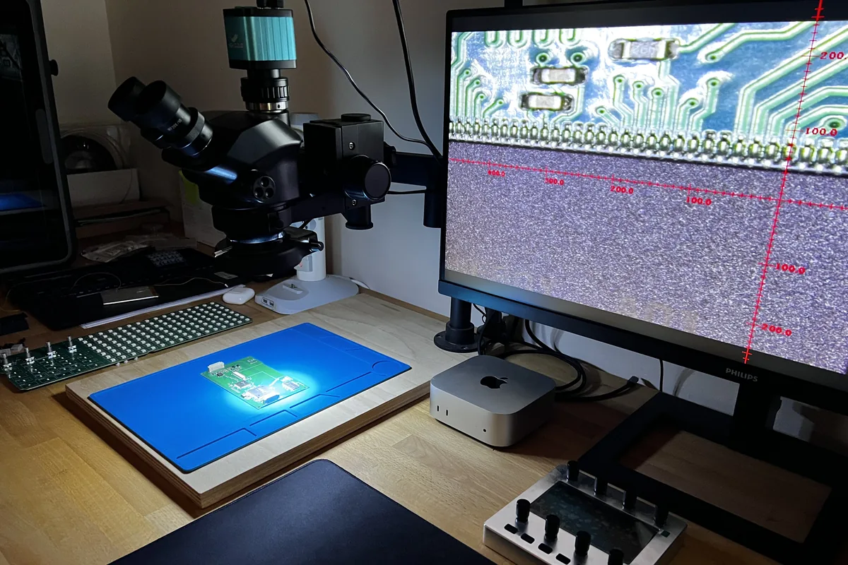 A circuit board on the inspection mat under a stereo microscope, magnified on the screen beside it